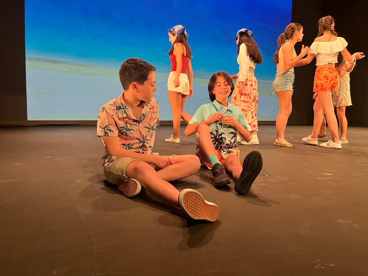 Ensayo en el escenario: dos alumnos sentados en el suelo y un grupo detrás.