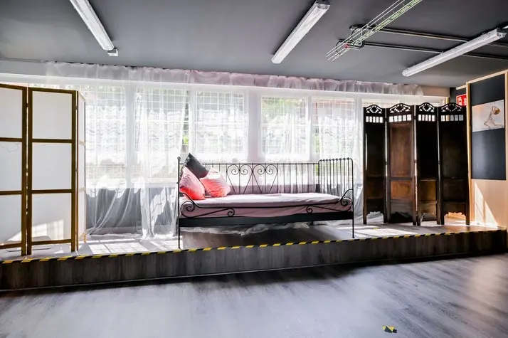 Tarima de escenario con cama de hierro, cojines y biombos de madera.