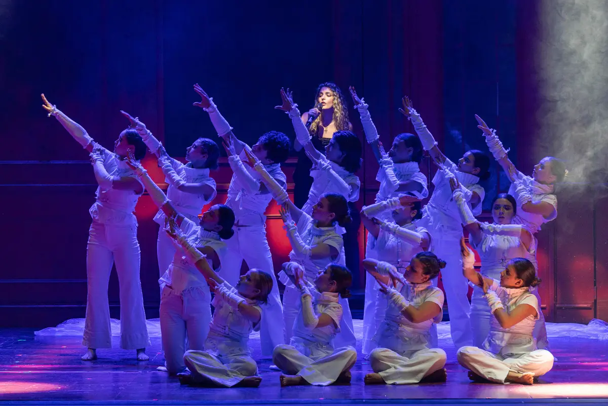 Un elenco vestido de blanco en el escenario, entre humo y luz violeta.